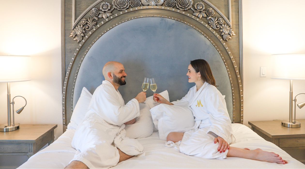 A man and a woman clink their wine glasses together as the sit facing one another smiling in a fancy bed wearing white robes at Mirbeau Inn and Spa in Rhinebeck